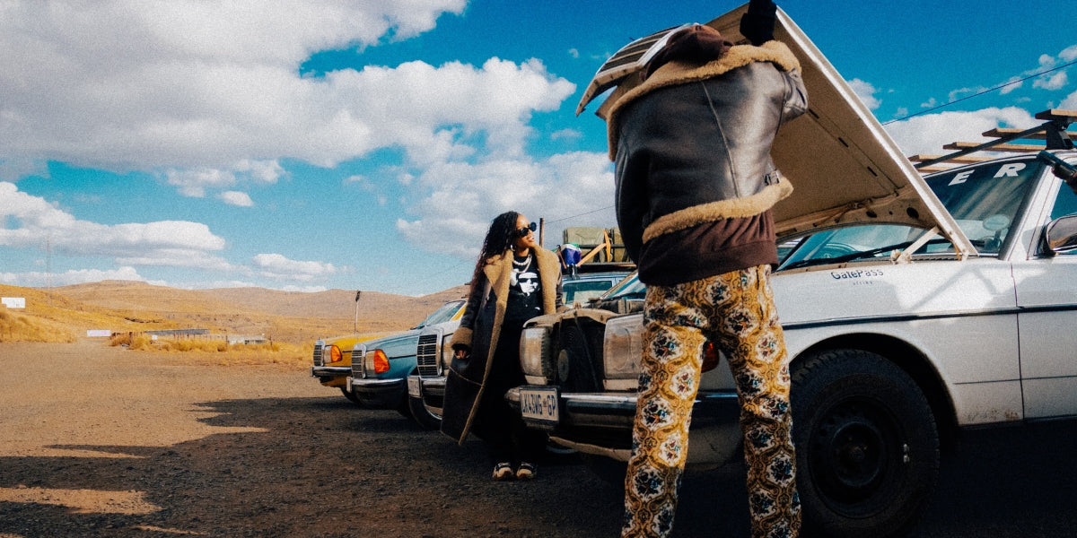 Street style fashion scene with two people in layered outfits standing by vintage cars in a desert landscape.